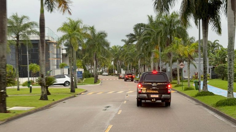 Viaturas da Polícia Federal circulam por uma rua arborizada.